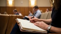 Professional woman taking detailed notes during corporate meeting or legal proceeding in modern conference room setting.