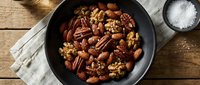 Mixed nuts including pecans and walnuts in black cast iron skillet on rustic wooden table with sea salt for healthy snacking and cooking ingredients.