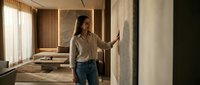 Young Asian woman architect examining concrete wall texture in modern minimalist interior with natural lighting and warm wood accents for construction planning.