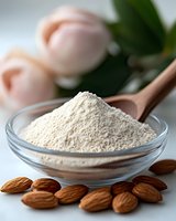 Ground almond flour in glass bowl with wooden spoon and raw almonds scattered on white surface, fresh tulips in soft focus background.