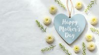 Blue heart-shaped sign with Happy Mother's Day text surrounded by yellow chrysanthemum flowers and green foliage on white marble background for greeting cards.