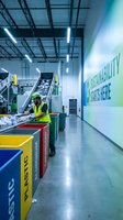 Workers in safety vests operating recycling sorting equipment in modern waste management facility with sustainability messaging on wall.