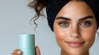 Young mixed race woman with curly hair and headwrap holding mint green cosmetic product container against light background, natural glowing makeup and nose piercing.