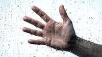Open palm of a person with dark skin tone pressing against wet glass surface covered with water droplets and condensation, conveying emotions of sadness, isolation, or despair.
