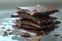 Broken dark chocolate pieces stacked in layers with crumbs scattered on light surface, macro food photography showing rich texture and glossy sheen.