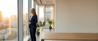 Professional businesswoman in dark suit standing by floor-to-ceiling windows in modern office space with wooden desk and city skyline view at sunset.