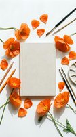 Blank canvas notebook surrounded by vibrant orange poppy flowers and art brushes on white background for creative projects and artistic inspiration.