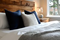 Cozy bedroom interior with rustic wooden headboard, navy blue and white pillows, textured bedding, and natural light from window.