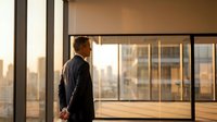 Confident Caucasian businessman in suit standing by office window at sunset, overlooking city skyline, reflecting on success and future vision.