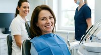 Middle-aged Caucasian woman smiling in dental chair during checkup with female dentist and male assistant in modern clinic office setting.