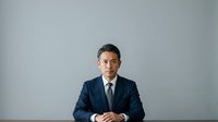 Asian male professional in dark business suit and tie seated against neutral gray background, suitable for corporate portraits, business websites, and professional headshots.