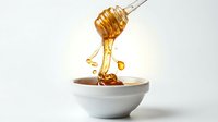 Golden honey dripping from a wooden dipper into a white ceramic bowl, isolated on white background, showcasing natural sweetness and organic food concept.