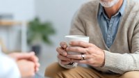 Senior man in beige cardigan holding white coffee cup during conversation in bright, modern living space with soft focus background.