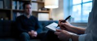 Professional therapist taking notes during counseling session with male patient in modern office setting for mental health treatment.