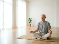 Senior man with light skin tone meditating in lotus position on yoga mat in bright, minimalist studio space with wooden floor and potted plant. Wellness and mindfulness practice.