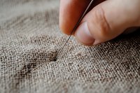 Close-up view of hand holding sewing needle piercing natural linen fabric, showing detailed texture and craftsmanship in soft, warm lighting and shallow depth of field.