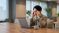 Young Asian businessman experiencing headache while working on laptop in modern office environment with coffee cup on wooden desk.