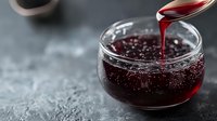 Red berry jam or jelly in glass bowl with spoon dripping sweet syrup, close up view on dark background, selective focus, food photography.