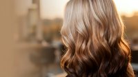 Young Caucasian woman with long wavy blonde hair styled in loose curls, backlit by warm golden sunlight creating beautiful highlights and depth.