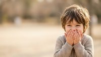 Young boy covering face with hands showing shy or embarrassed expression outdoors in natural lighting for childhood emotion concepts.