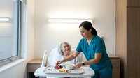 Healthcare worker serving meal to elderly patient in hospital room with natural lighting and modern medical facility interior.