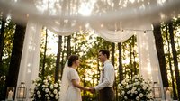 Romantic outdoor wedding ceremony with bride and groom exchanging vows under illuminated fabric canopy surrounded by lush greenery and floral arrangements.