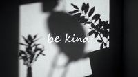 Monochrome shadow play on wall with plant silhouettes and handwritten be kind motivational quote in elegant script.