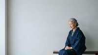 Senior Asian woman with gray hair wearing dark blue kimono-style robe, sitting contemplatively against white wall. Suitable for wellness, mindfulness, aging gracefully, and lifestyle editorial use.