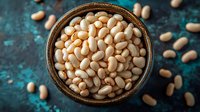White navy beans in rustic wooden bowl on dark turquoise background with scattered legumes. Fresh organic protein rich food for healthy eating and cooking.