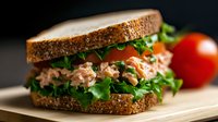 Fresh tuna salad sandwich with crisp lettuce on whole grain bread, served with ripe tomato on wooden cutting board against dark background.