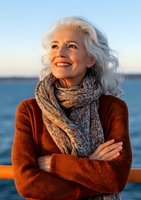 Confident mature woman with silver hair smiling at sunset, wearing brown sweater and patterned scarf, standing by ocean with crossed arms and natural makeup.