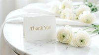Elegant thank you card with gold lettering beside fresh white ranunculus flowers on a marble table, soft light, wedding or event stationery concept.