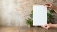 Hands holding blank white paper sheet against brick wall background with green plants, mockup for branding, advertising, and design presentations.