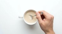 Caucasian male hand stirring creamy soup or sauce in white ceramic bowl with spoon on white background, overhead view for cooking and culinary content.
