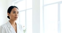 Young woman with dark hair and South Asian ethnicity wearing white lab coat in bright modern office setting, suitable for healthcare, medical, and professional business concepts.