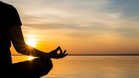Person meditating in lotus pose by calm water during golden sunset with peaceful sky reflection creating serene wellness atmosphere for mindfulness practice.