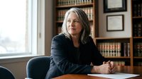 Mature woman with long gray hair and fair complexion, wearing dark blazer, seated at desk in professional office setting with bookshelves and framed certificates behind her, suitable for corporate, legal, or executive business concepts.