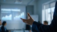 Businessman in dark suit gesturing during corporate presentation with analytics charts displayed on screen behind him in modern office conference room setting.