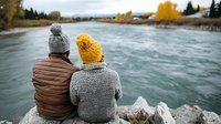 Couple in warm winter clothing sitting on rocks, enjoying scenic river view with autumn trees and mountains in background.