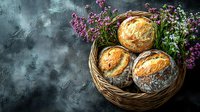 Fresh crusty artisan bread loaves in rustic wicker basket with purple wildflowers against dark moody background, food photography styling for bakery.