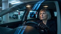 Middle-aged Caucasian woman with blonde bob haircut driving car in underground parking garage with focused expression and interior lighting.
