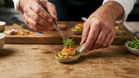 Chef plating gourmet appetizer with microgreens on wooden surface, professional food preparation and culinary presentation for restaurants.