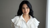 Young woman with long black hair and South Asian ethnicity wearing white ruffled collar blouse, professional headshot against neutral gray background for corporate, business, and portrait use.