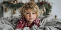 Young child with curly blonde hair and blue eyes wearing red patterned pajamas, lying on gray bedding during holiday season with festive decorations in background.
