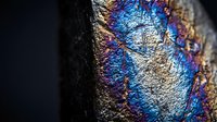 Iridescent metallic surface with rainbow oil slick colors showing blue purple gold reflections on textured material for abstract backgrounds and design projects.
