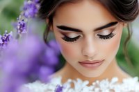 Young bride with elegant makeup and long eyelashes surrounded by purple flowers in natural outdoor wedding setting with soft romantic lighting.