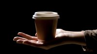 Hand holding brown disposable coffee cup with lid against dark background, morning beverage service concept for cafe and restaurant business marketing.
