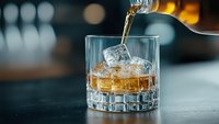 Whiskey being poured into crystal glass with ice cubes on dark bar counter. Premium spirits, cocktail preparation, luxury drinking experience.