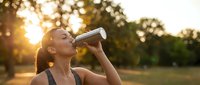 Young athletic woman drinking water from bottle during outdoor workout at golden hour sunset for hydration and fitness recovery.