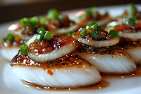 Fresh steamed fish fillets topped with spicy chili sauce, sliced green onions and sauteed garlic in Asian style marinade, served on white plate, macro food photography.